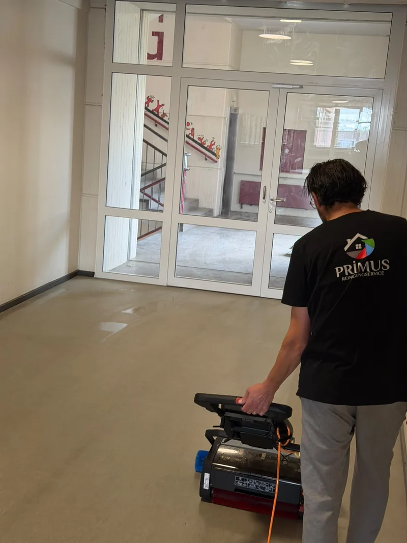 Person mit Primus-Shirt und Bodenreinigungsmaschine vor einer Glastür.